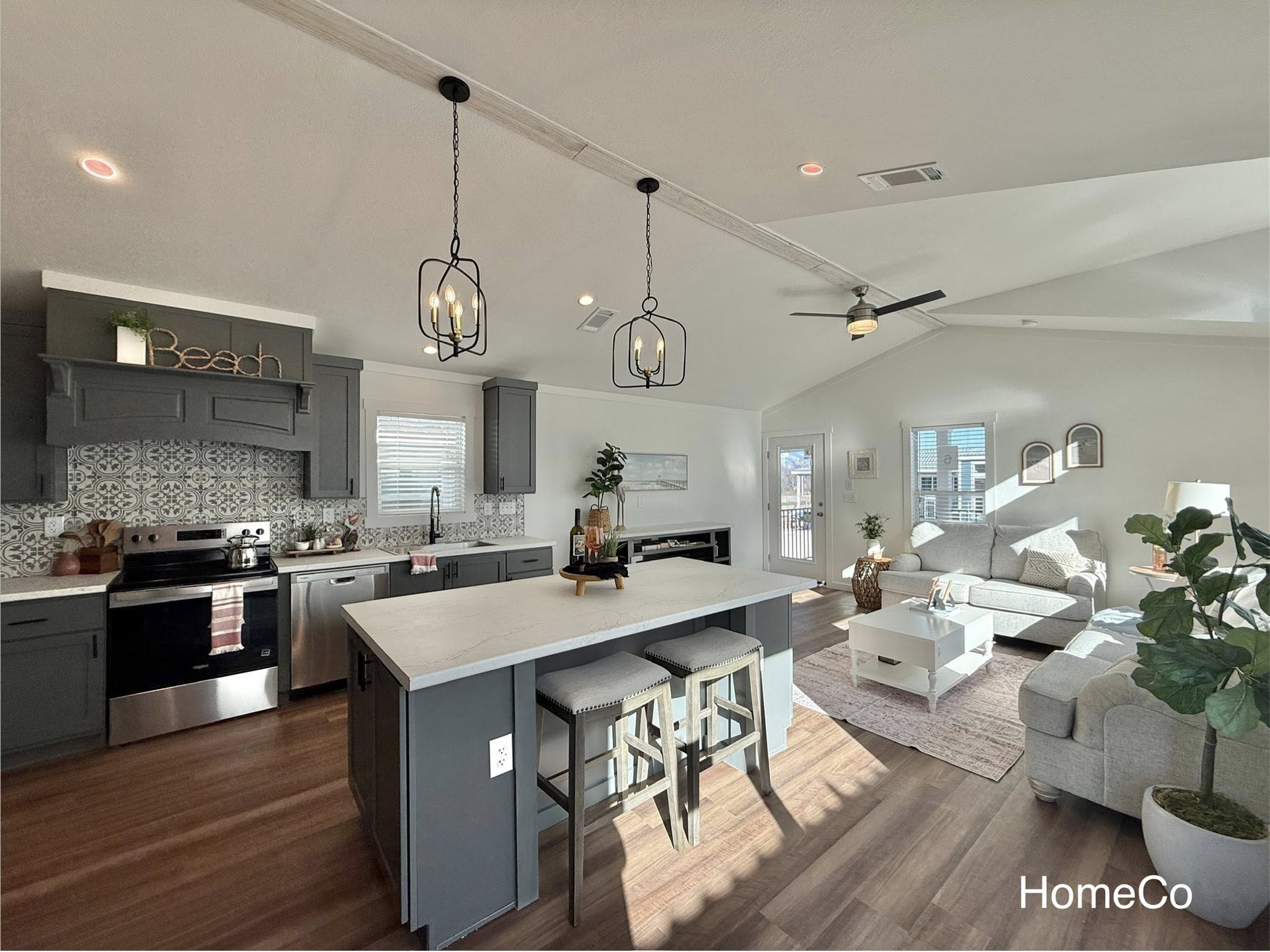 Bright, modern open-plan kitchen and living room. Features dark cabinets, light countertops, a large island with stools, and a cozy seating area. Warm and inviting decor.