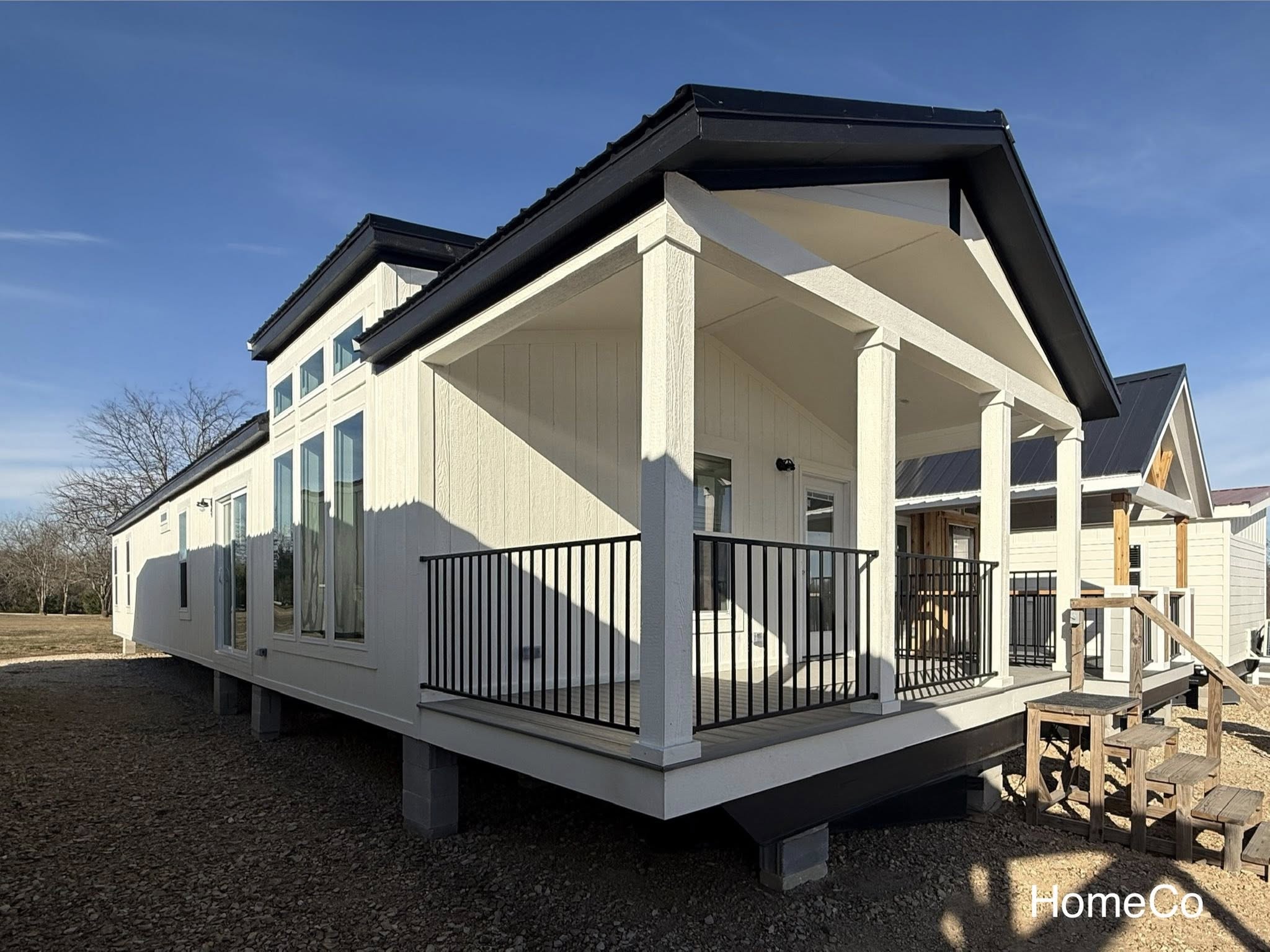 A modern white manufactured home with a black roof and large windows. The home features a small porch with metal railings and wooden steps.