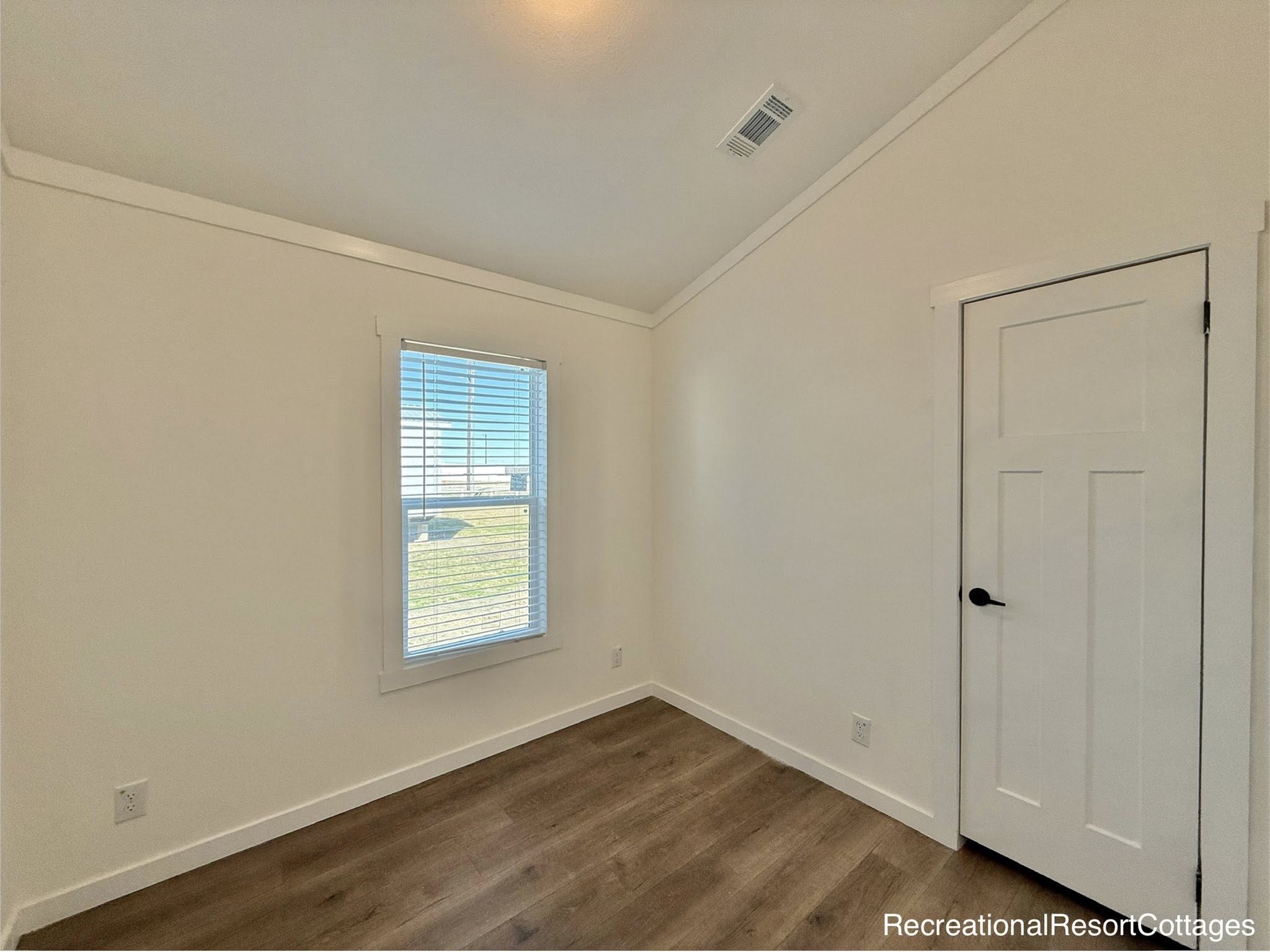 Minimalist room with white walls, wood floor, and a single window with blinds. A white door with a black handle stands on the right. Bright and airy ambiance.