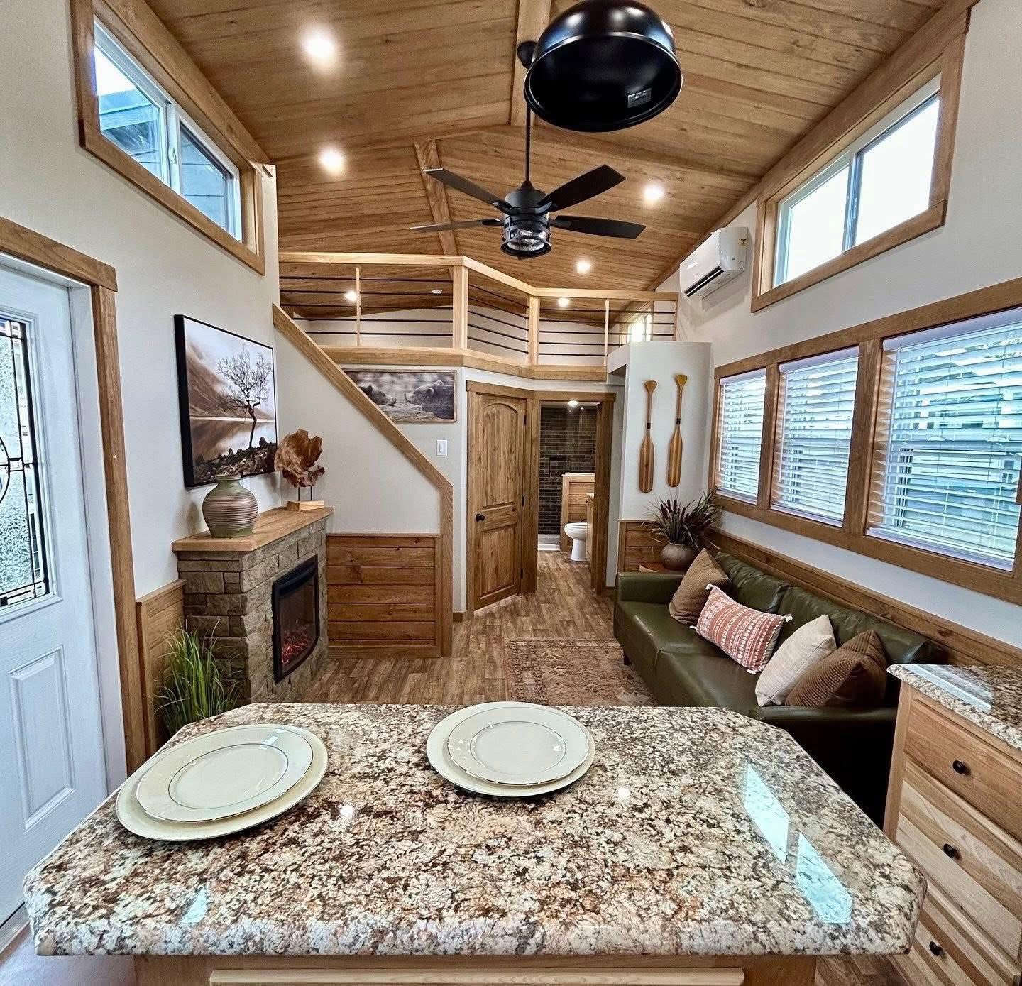 Cozy tiny house interior with wood accents features a kitchen island, green sofa, stone fireplace, loft, ceiling fan, and large windows.