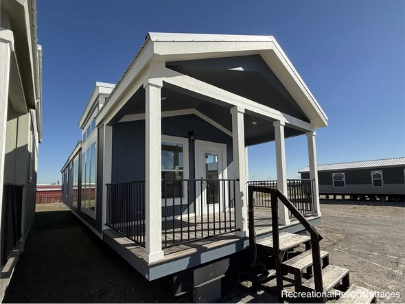 Small blue mobile home with a white-trimmed porch, stairs, and black railing. It’s set on a dirt plot under a clear blue sky, evoking a clean, modern feel.