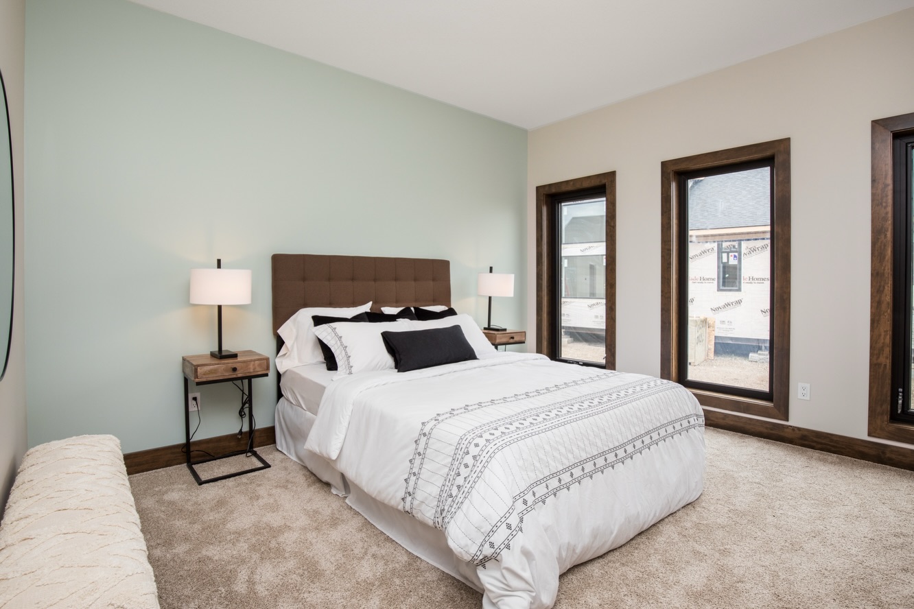 Modern bedroom with light green accent wall, brown headboard, and a white bedspread. Flanked by two lamps and large windows, the space feels airy and serene.