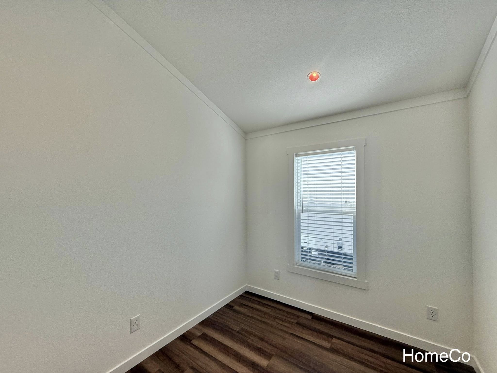 Small, empty room with white walls and dark wooden floor. A single window with blinds lets in natural light. A recessed ceiling light is on.