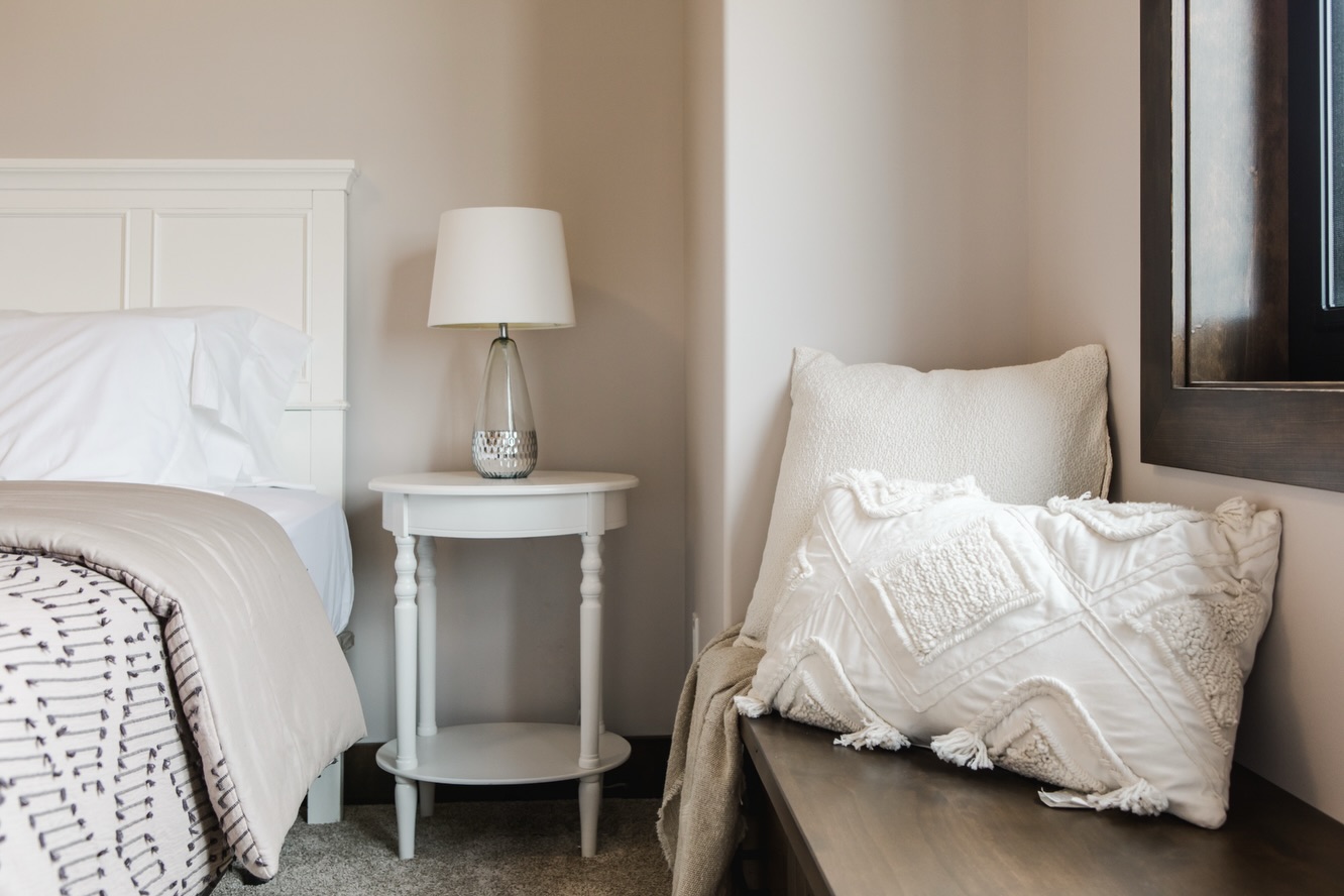 Cozy bedroom corner with a white bed, a beige blanket, an elegant side table with a lamp, and textured pillows on a bench. Calm, inviting ambiance.