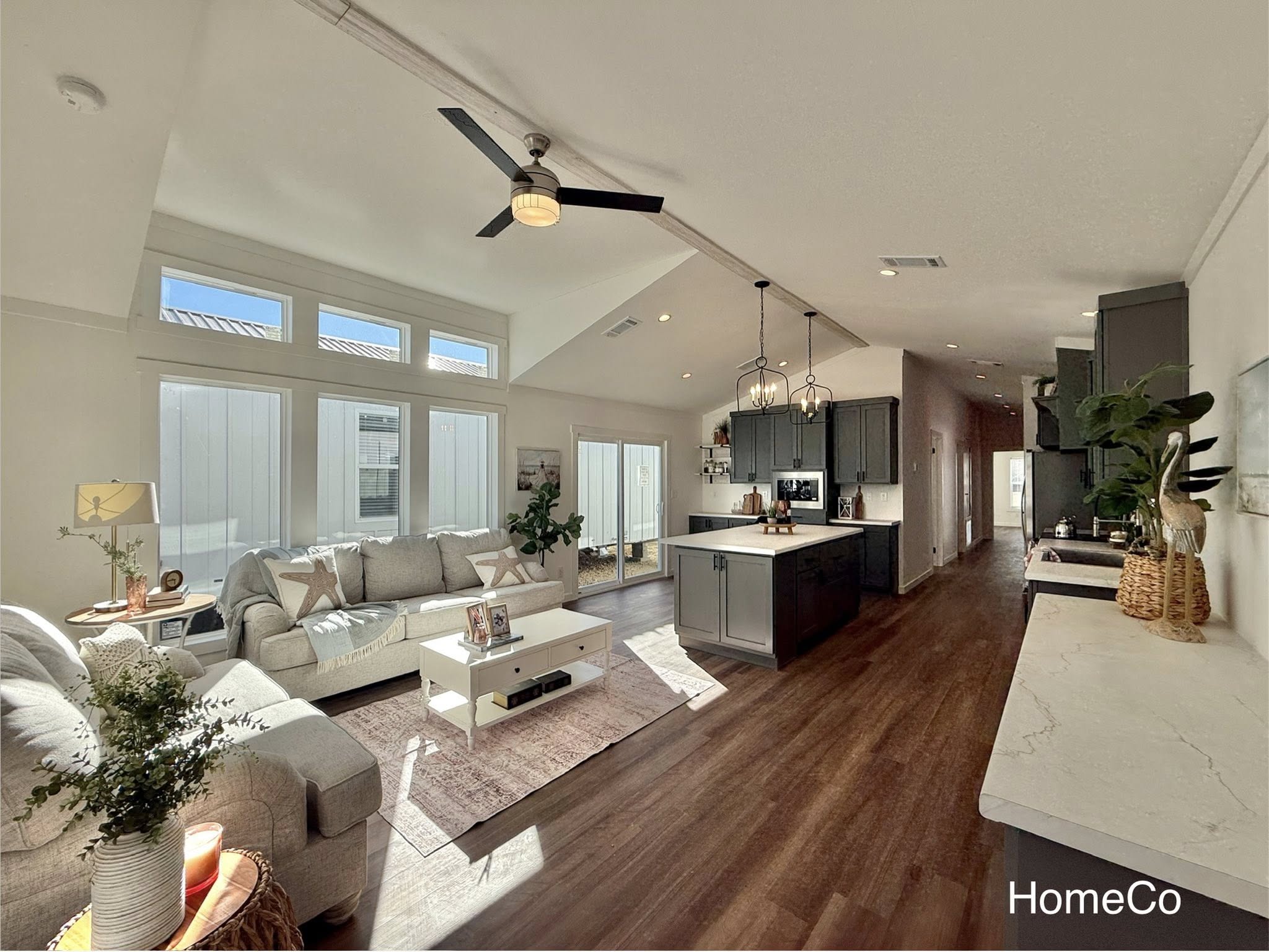 Bright, modern open-plan living room and kitchen. Features large windows, gray sectional sofa, white coffee table, kitchen island, and warm wood floors. Sleek, inviting atmosphere.