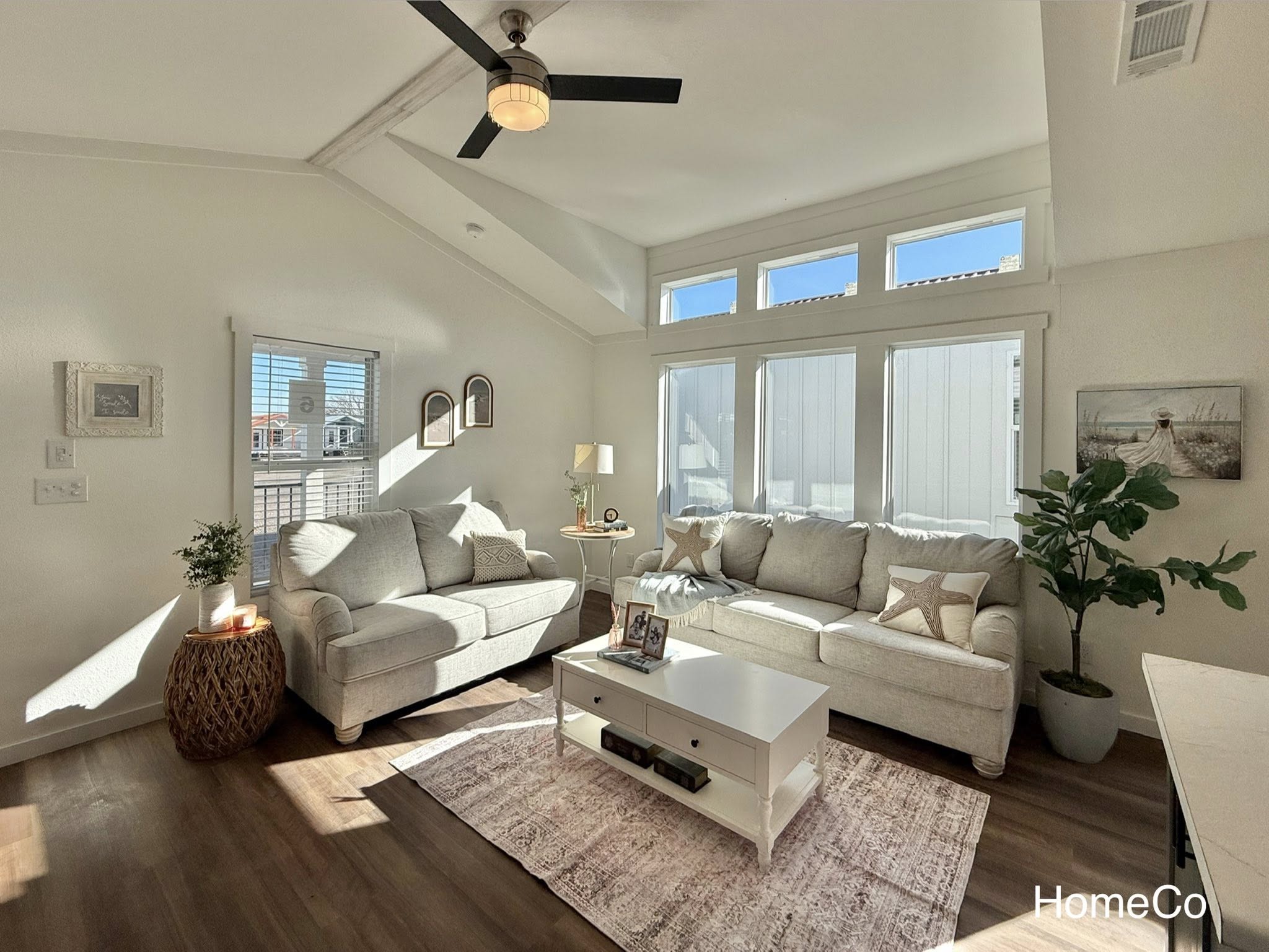 Bright living room with two beige sofas, sunlight streaming through large windows. Modern decor includes a ceiling fan, white coffee table, and potted plant. Cozy ambiance.