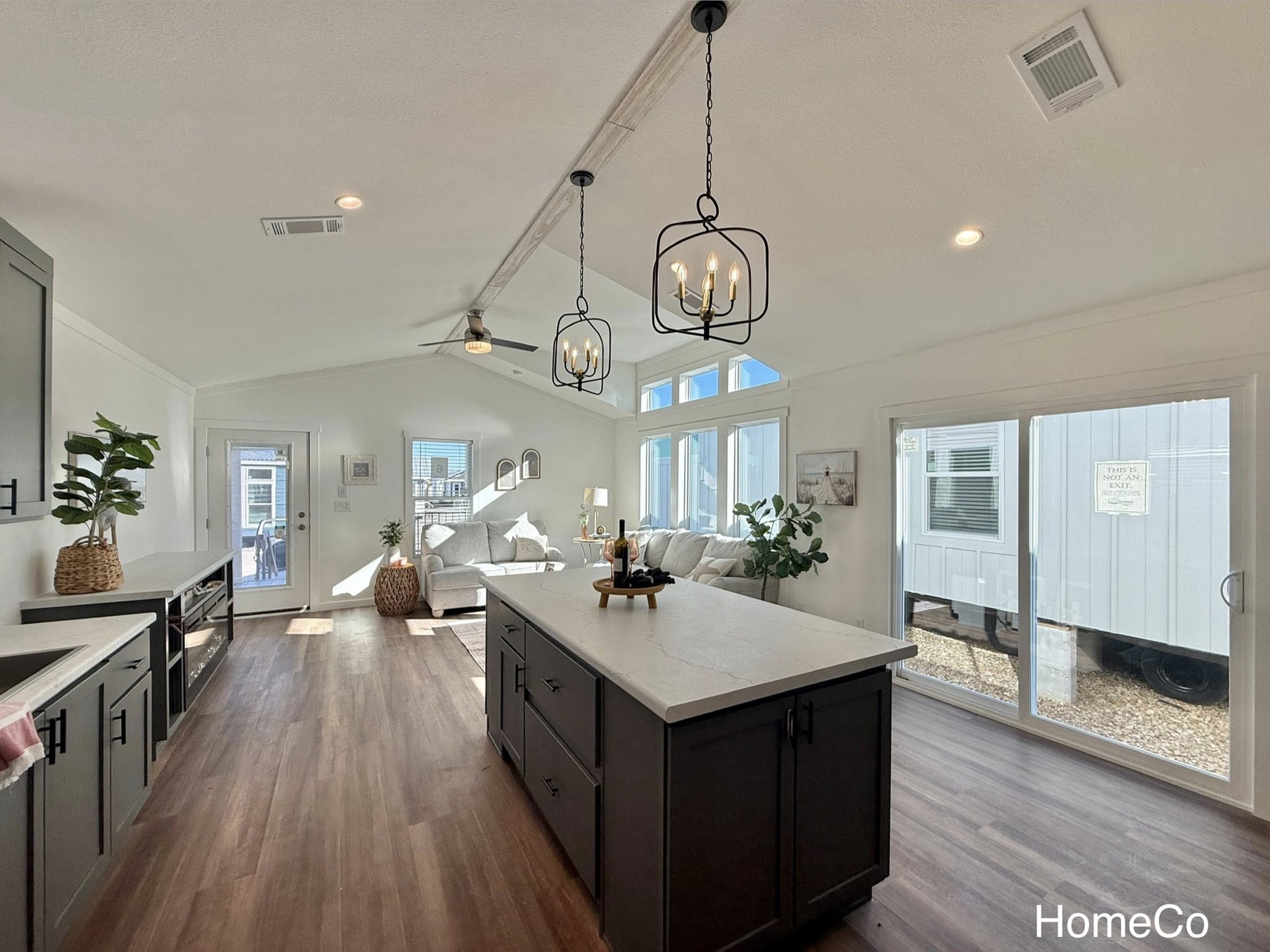 Modern, sunlit kitchen and living area with dark cabinetry, white island, and wood flooring. Large windows, cozy beige sofa, and hanging lights.