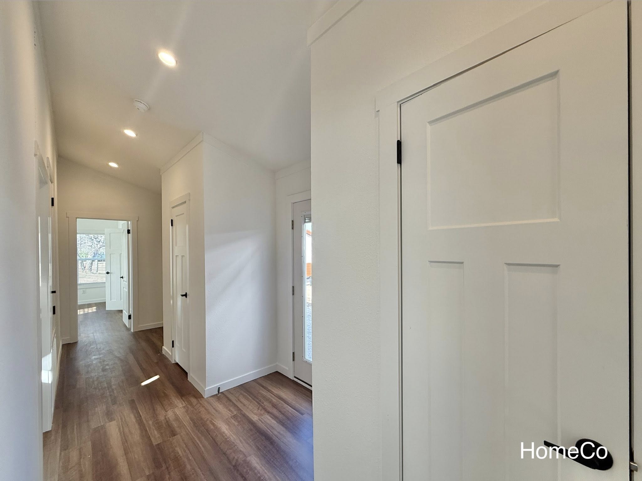 Modern hallway with wooden floors and white walls, featuring multiple doors. Natural light from a window at the end creates an airy atmosphere.