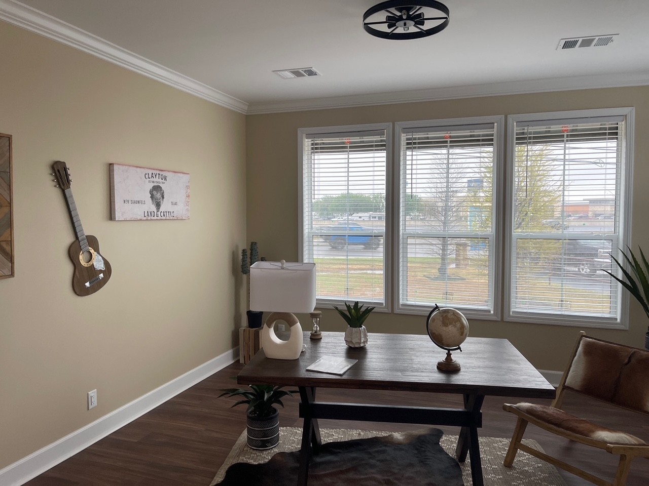 A cozy office with a wooden desk, globe, and lamp. A guitar and painting decorate the beige walls. Large windows let in natural light, creating a warm atmosphere.