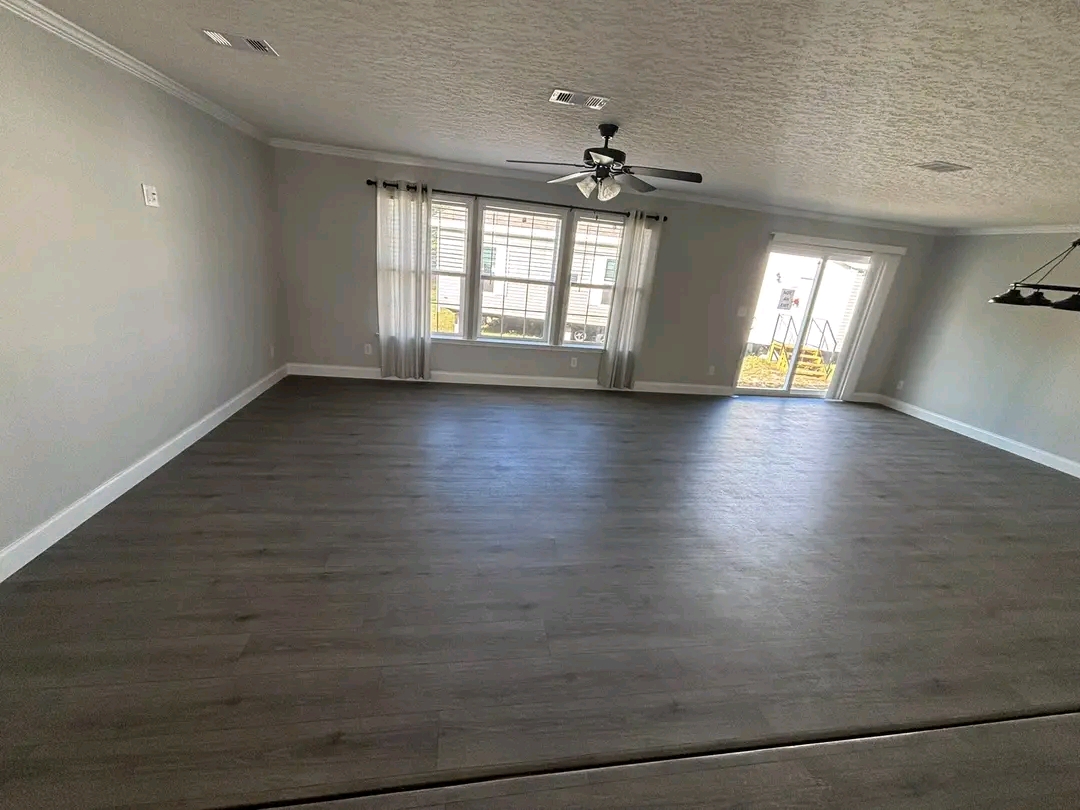 Spacious empty room with gray walls and dark hardwood floors. Large windows with sheer curtains and a ceiling fan create a bright, airy atmosphere.