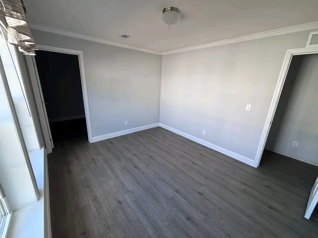 An empty room with light gray walls, a ceiling light, and wood flooring. Two doorways lead to other rooms, and a window brings in natural light.