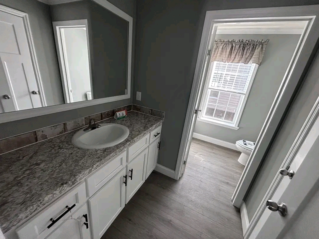 Modern bathroom with white cabinets, granite countertop, and mirror. Gray walls, open door to room with window, light wood floor, calm ambiance.
