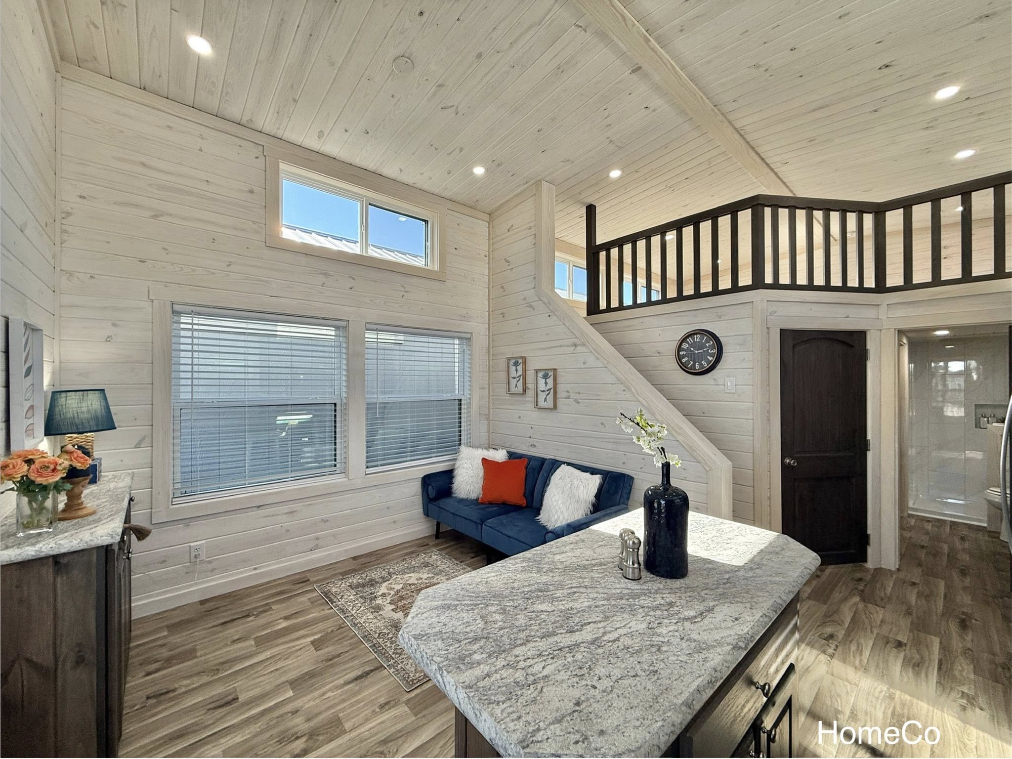 Modern tiny home interior with light wood paneling, a loft, and high windows. Blue sofa with bright cushions, granite countertop, and cozy decor.