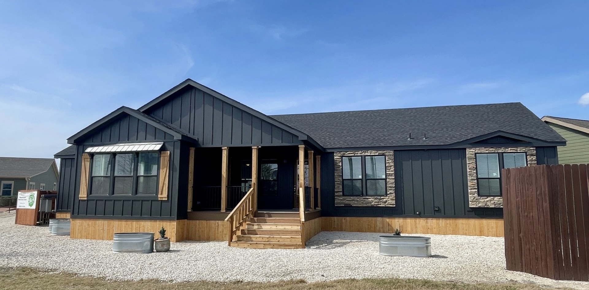 Single-story dark gray house with a gabled roof, stone accents, and wooden steps. It has large windows, wooden trim, and a landscaped front yard.