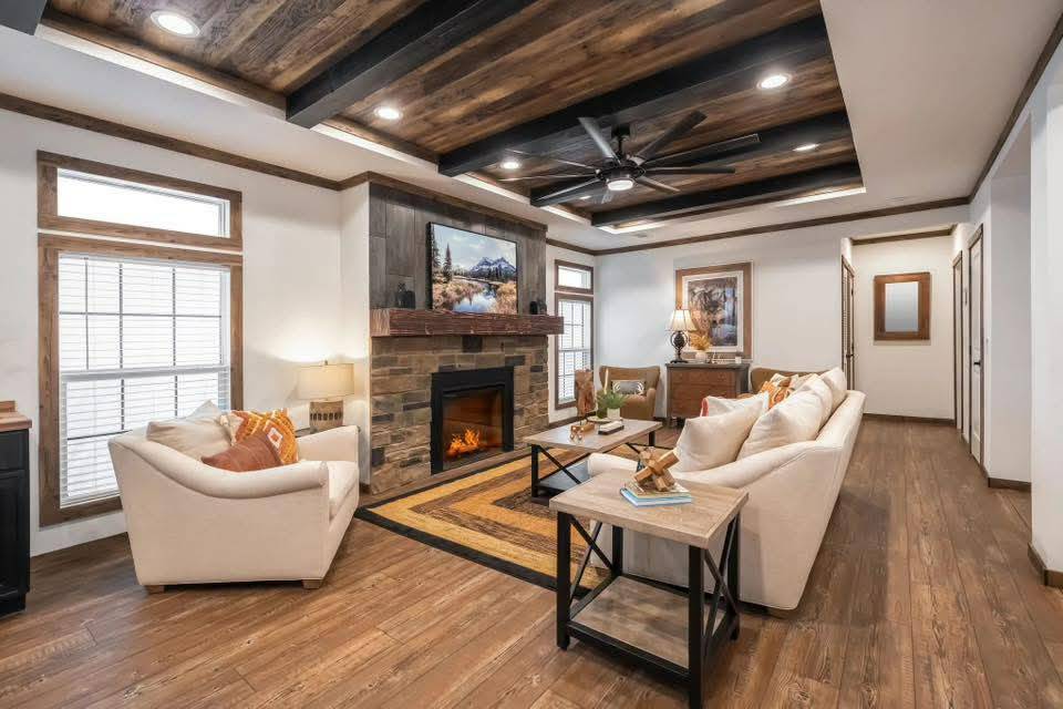 Warm, cozy living room with wooden floors and ceiling beams. Beige sofas, stone fireplace, and a ceiling fan create a rustic, inviting ambiance.