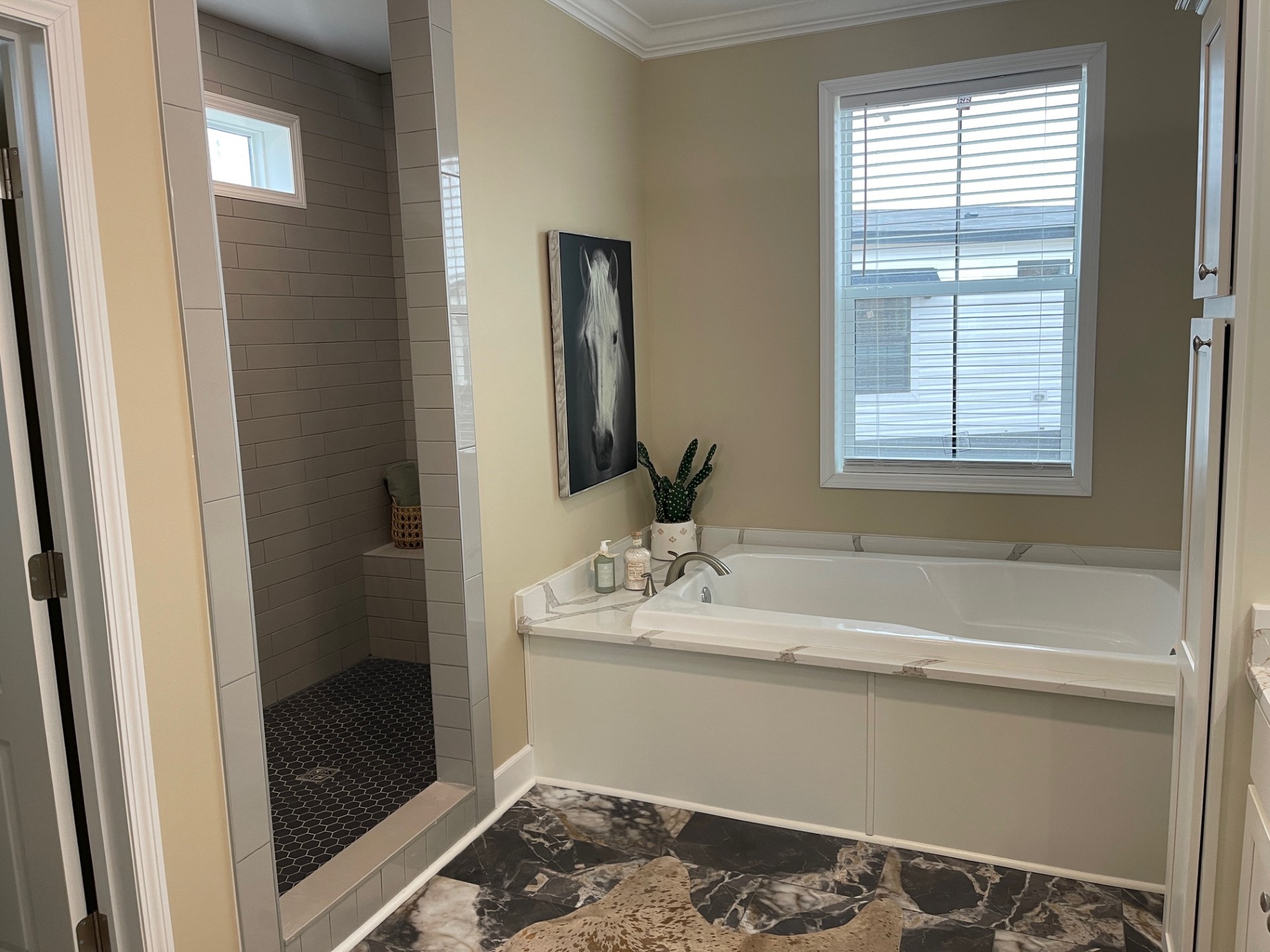 Modern bathroom featuring a large bathtub under a window with blinds, a tiled walk-in shower, and a horse painting. The space feels serene and elegant.