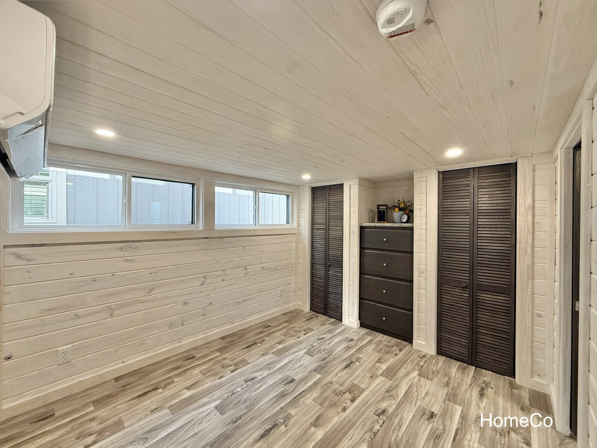 Cozy room with white wooden walls and ceiling, wood laminate flooring, two dark brown closets, and a black dresser. Bright windows add a fresh feel.