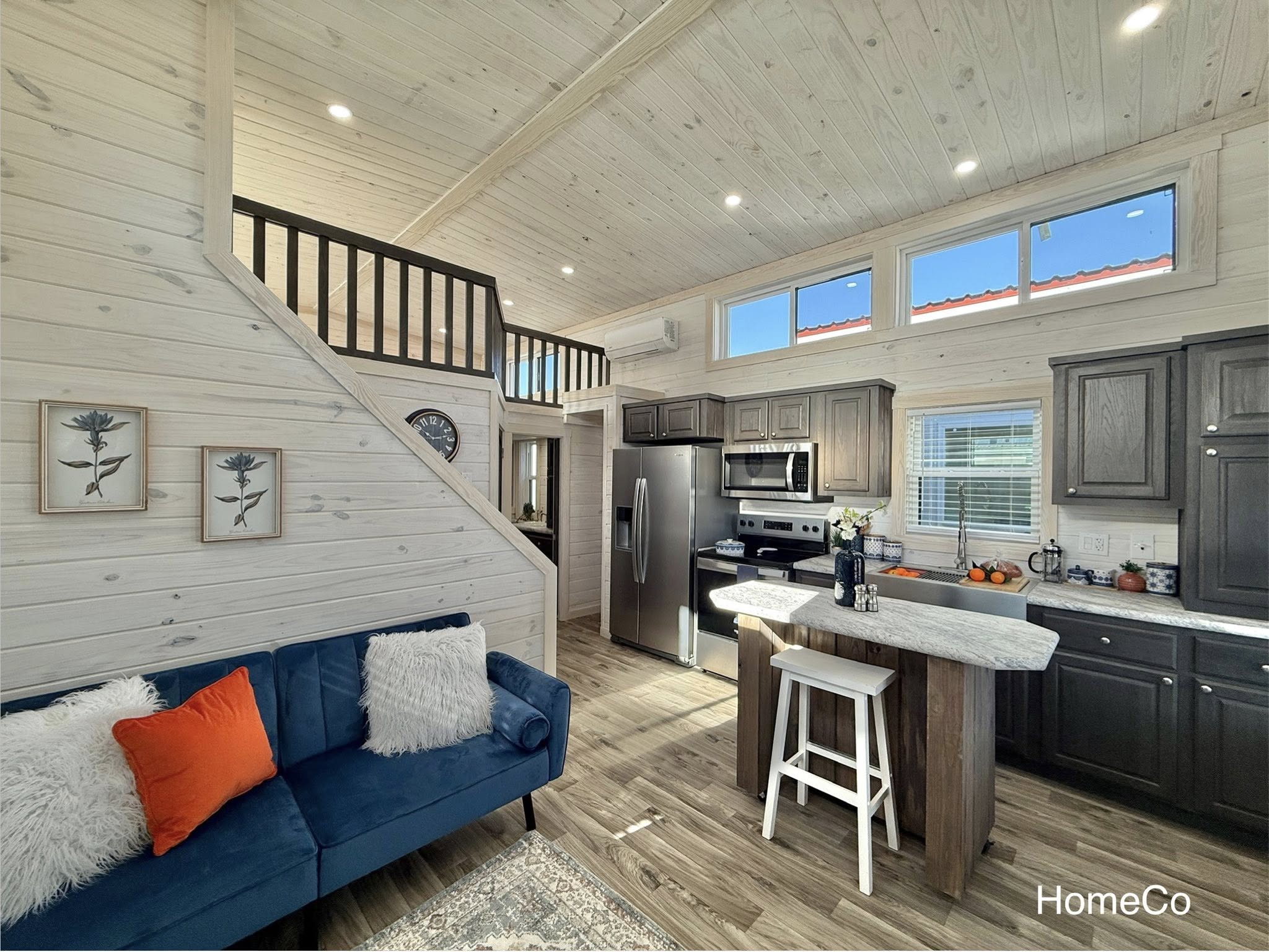 Cozy modern living space with a loft. Features a blue sofa, a compact kitchen with stainless appliances, and warm wood accents, creating an inviting atmosphere.