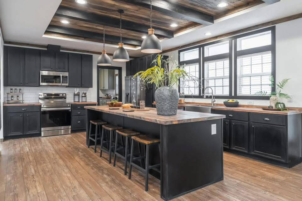 Spacious modern kitchen with black cabinets, wooden countertops, and an island with stools. Pendant lights hang above, and large windows brighten the space.