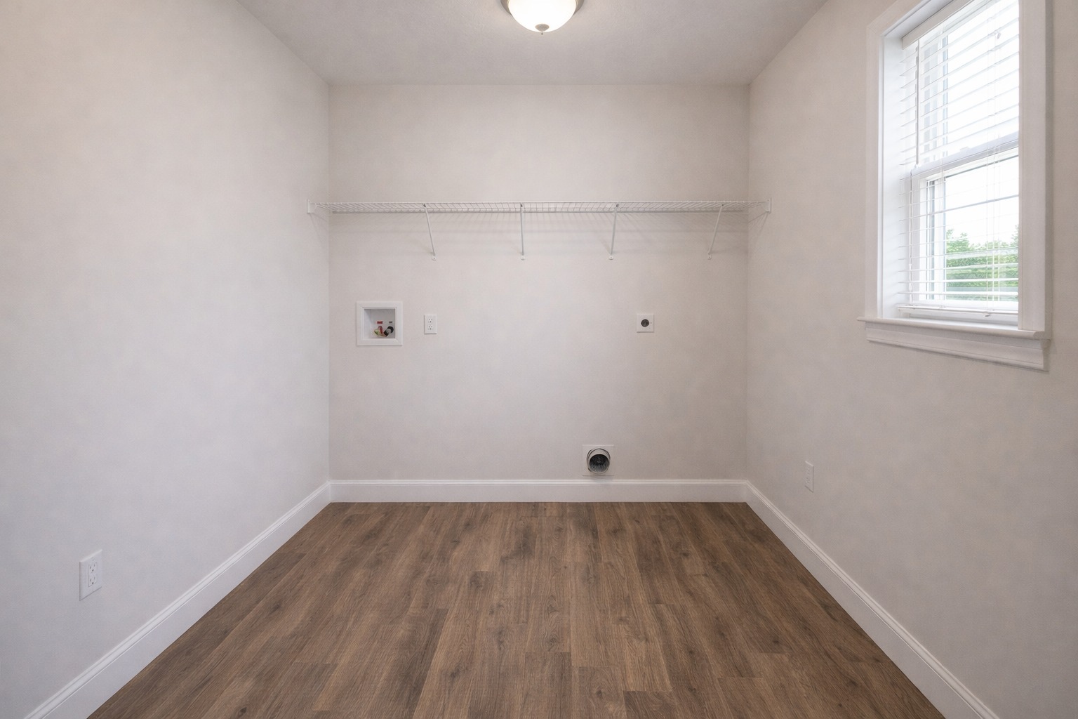Empty laundry room with light gray walls, wood floor, and large window on the right. A wire shelf spans the back wall, and utility outlets are visible. Bright and minimal.
