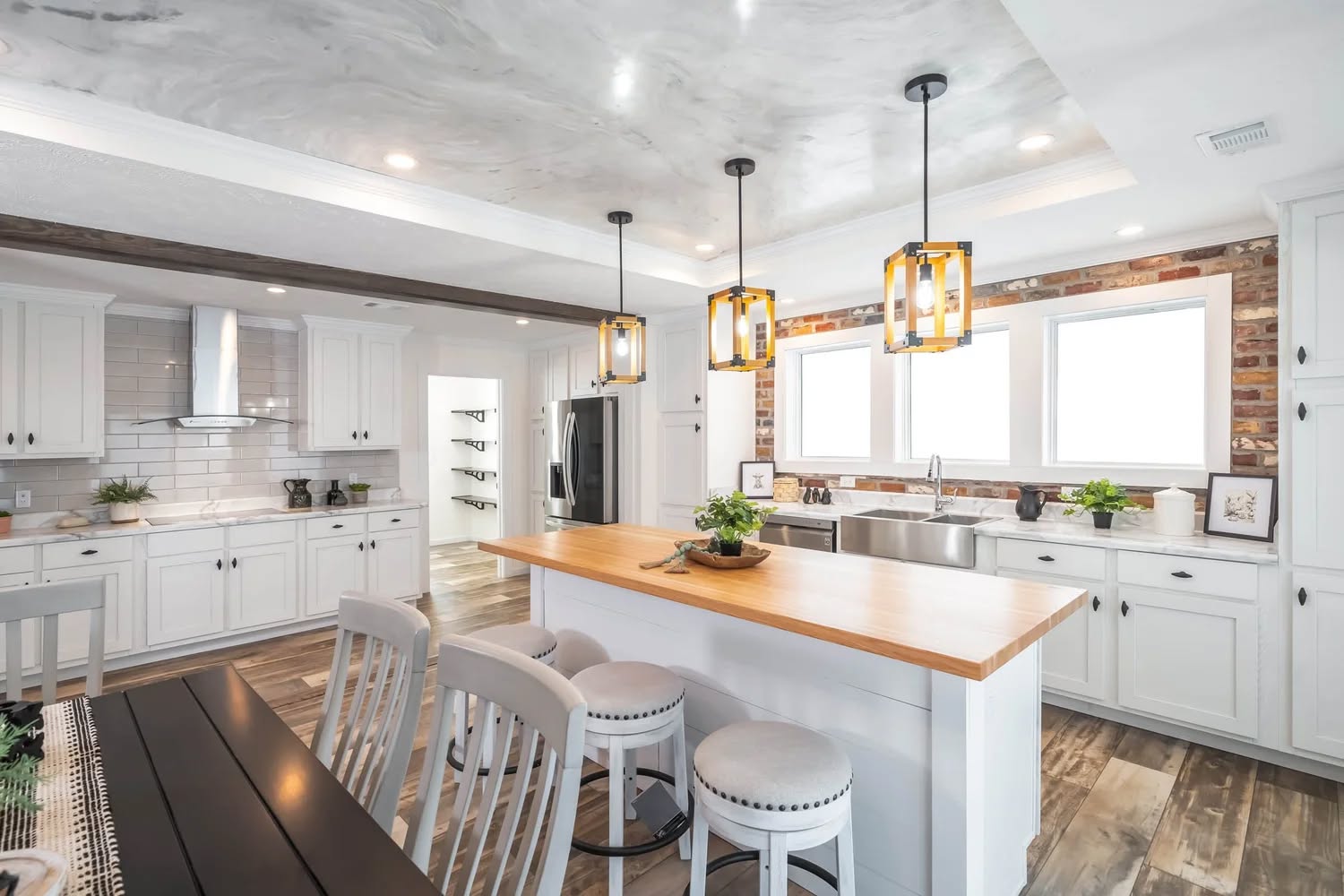 Spacious kitchen with white cabinets, wooden island, and modern pendant lights. Three stools, large windows, and plants create a bright, welcoming atmosphere.