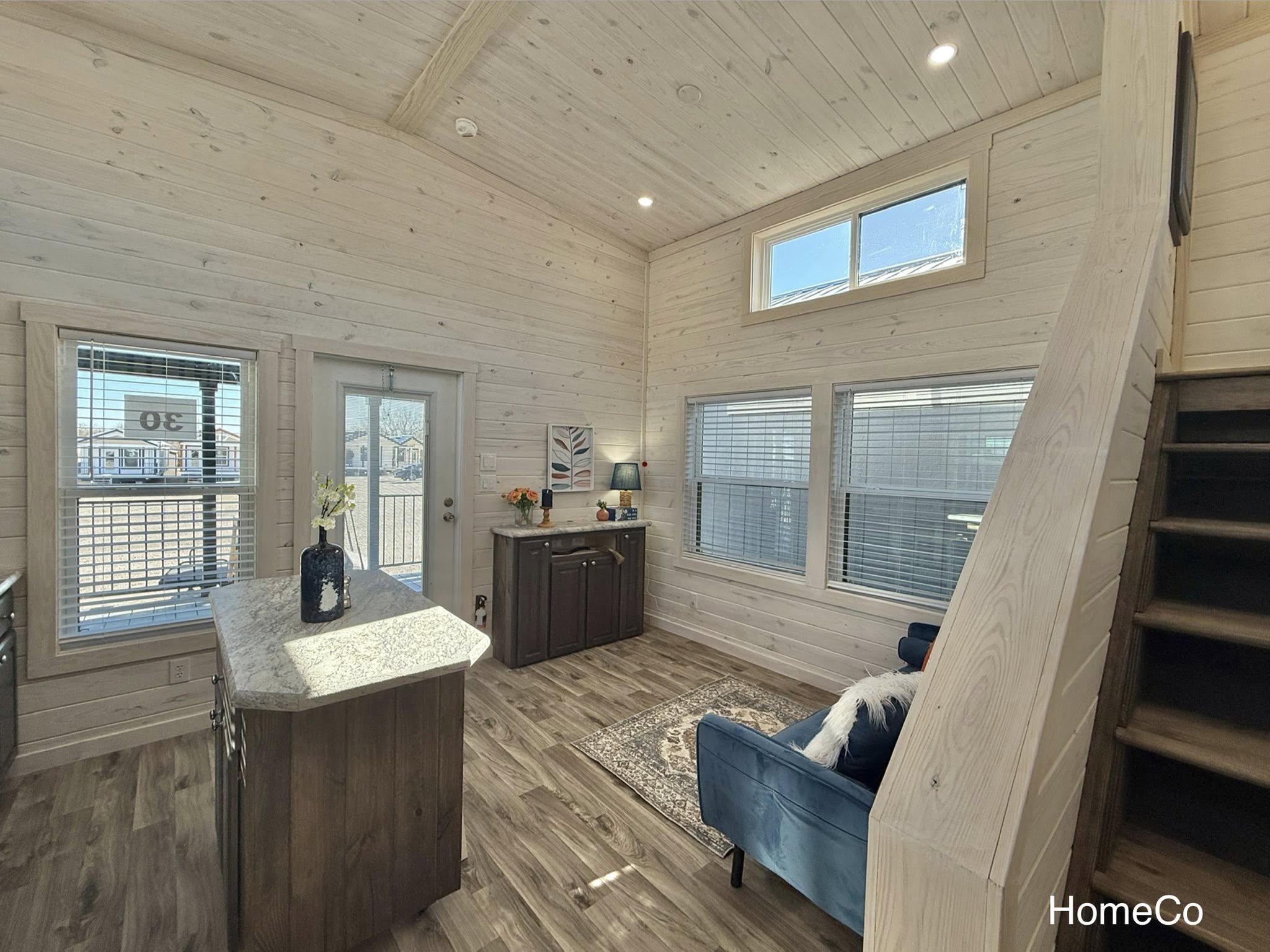 Cozy living room with wooden walls and floors, featuring a navy blue armchair, small kitchen island, windows with blinds, and a staircase. Bright, welcoming atmosphere.