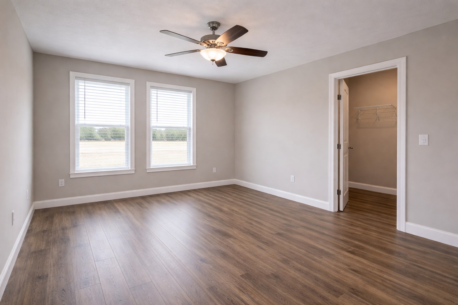 An empty room with light gray walls, two large windows, and wooden floors. A ceiling fan with lights sits above, creating a bright, spacious feel.