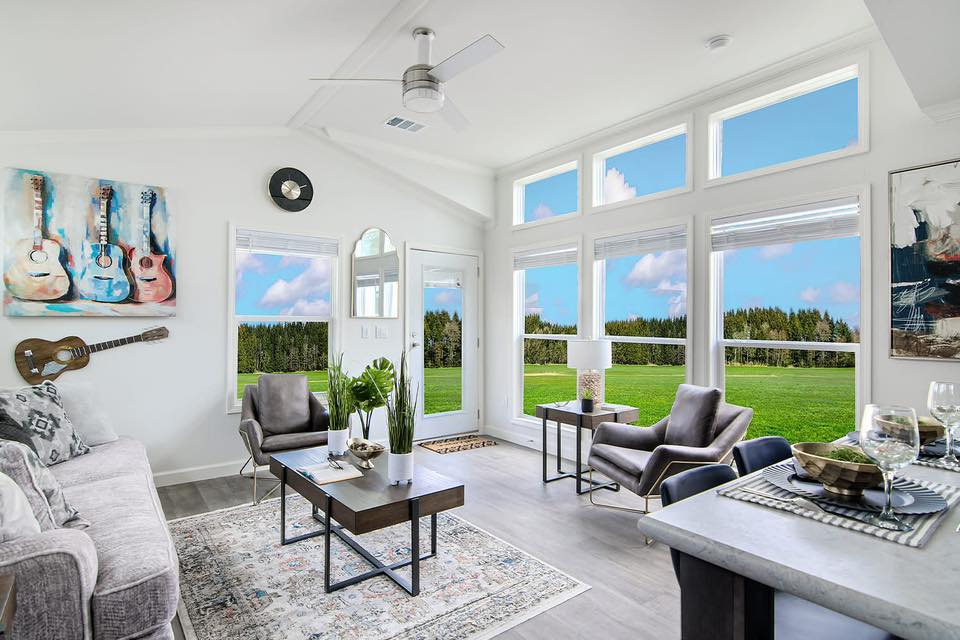 Bright, modern living room with large windows showcasing a scenic view of a green field and blue sky. Neutral furnishings, guitar art, and elegant decor.