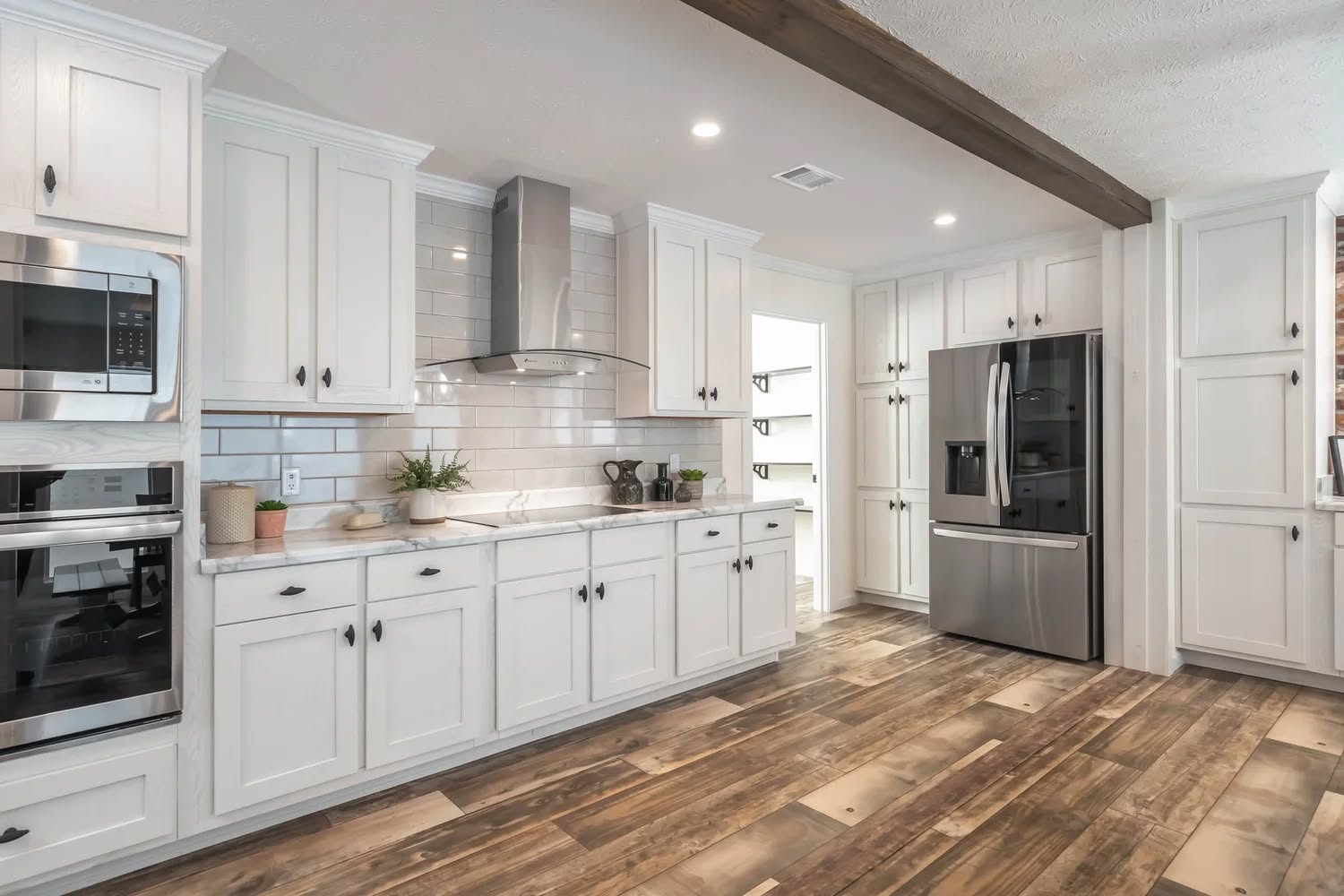 A modern kitchen with white cabinets, stainless steel appliances, and wooden floors. The bright space features subway tiles and a decorative beam.