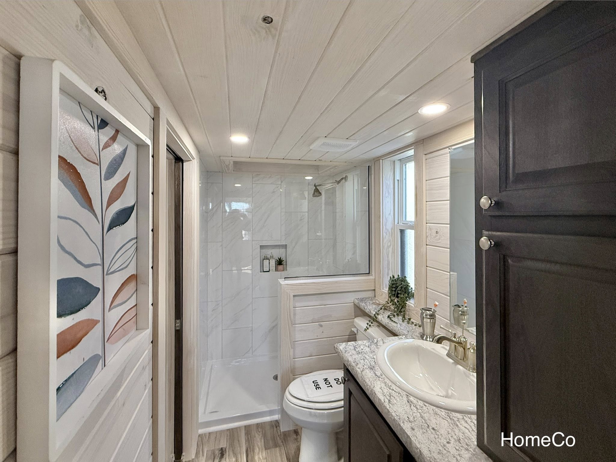 Bright, modern bathroom with wood paneling, white tile shower, granite countertop, and dark cabinets. Minimalist art adds a stylish touch.
