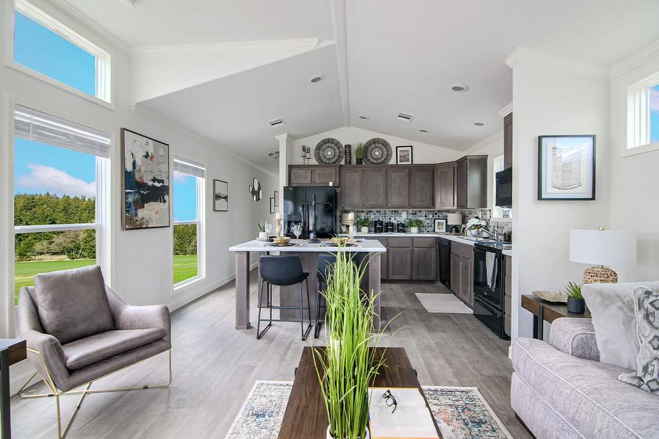 Modern open-plan living space with a cozy neutral palette. Large windows reveal a lush green view. The stylish kitchen island has chic bar stools, and the room is accented with abstract art and greenery, creating a serene, inviting atmosphere.