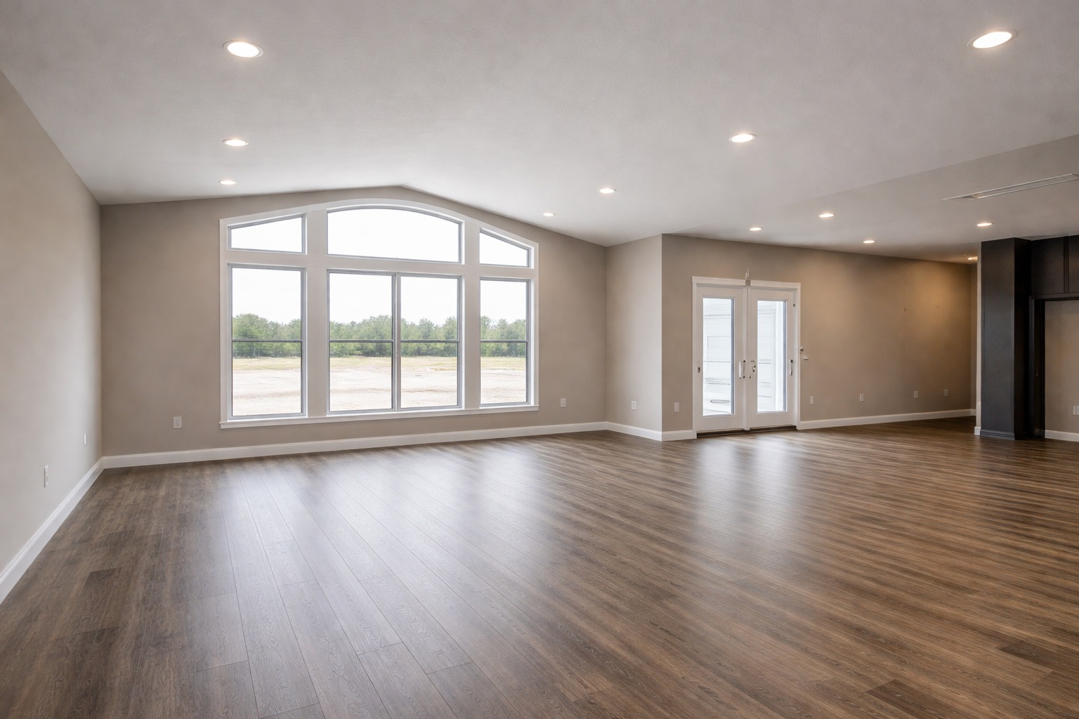Spacious, empty room with large arched window and double doors. Hardwood floors, beige walls, and recessed lighting create a bright, open atmosphere.