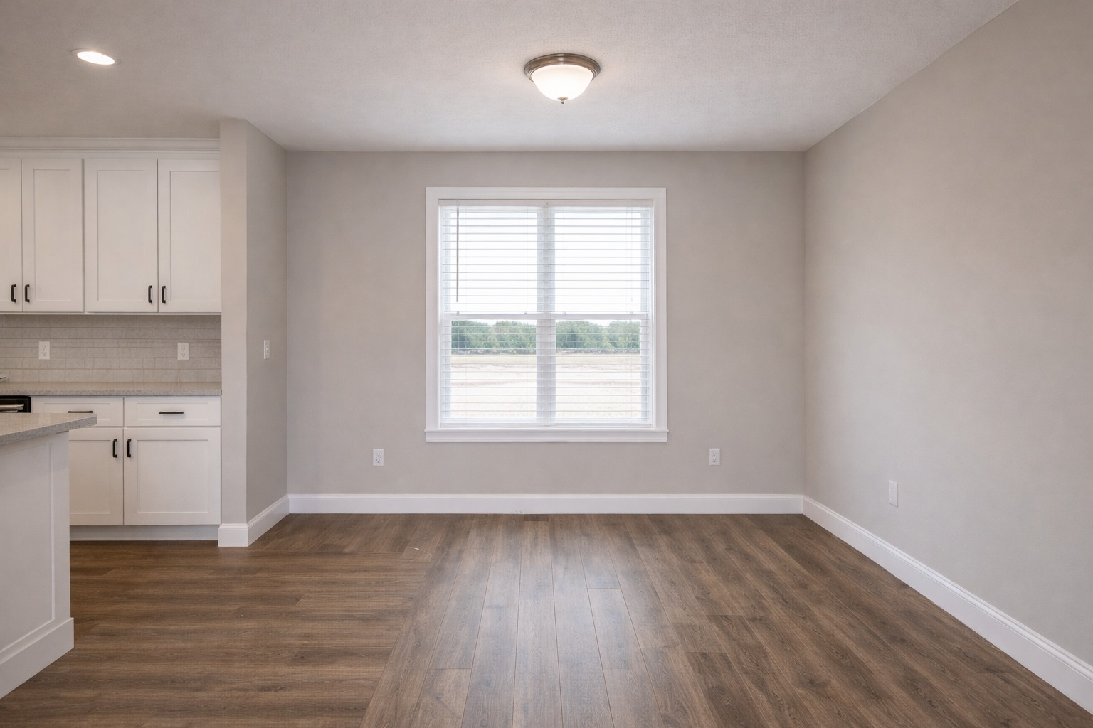 Minimalist empty room featuring light gray walls, wooden floor, and a window with white blinds. White kitchen cabinets on the left add a modern touch.