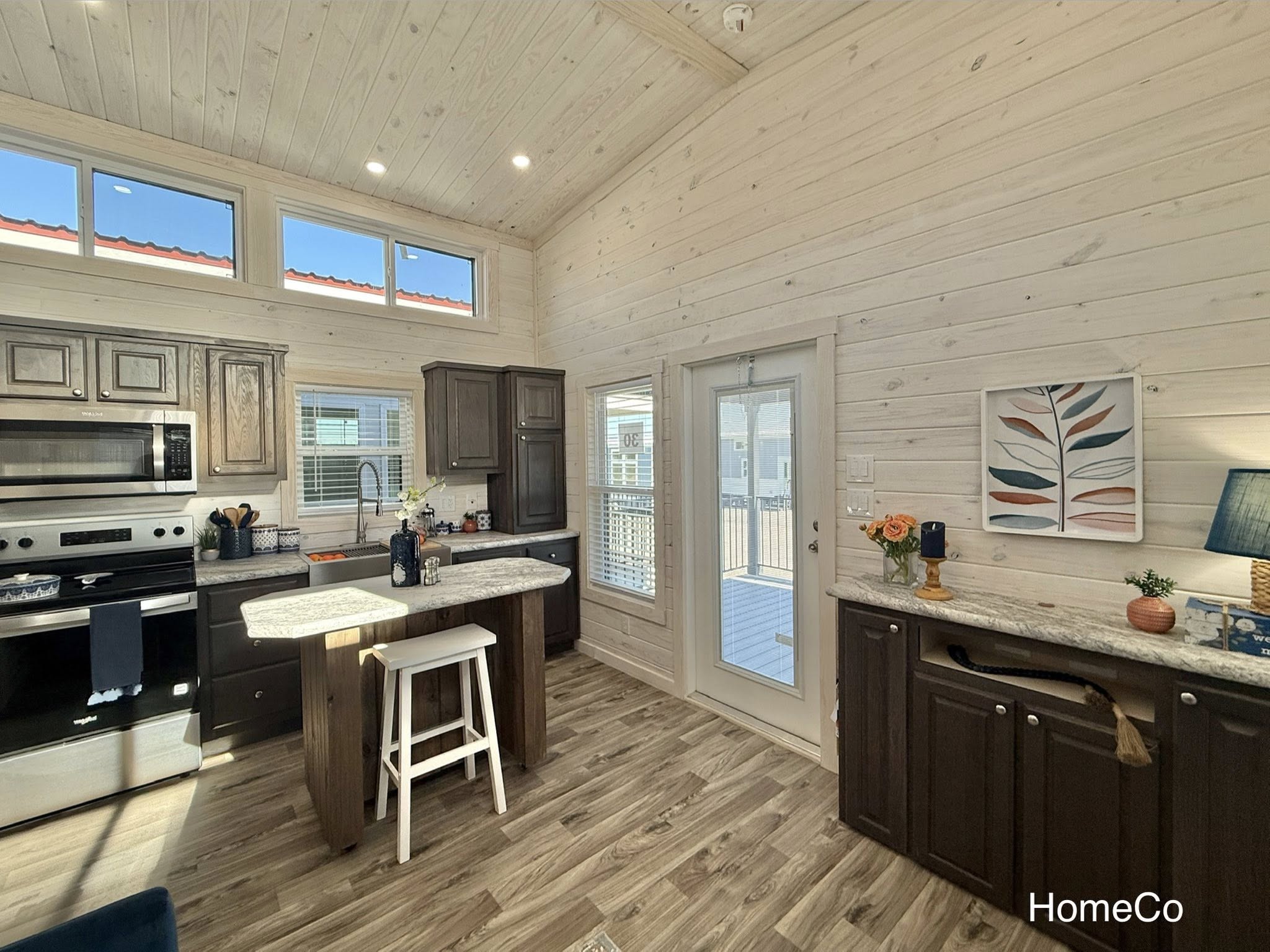 Bright kitchen with wood-paneled walls and ceiling, modern appliances, a central island with a stool, and a door leading outside. Cozy and inviting.