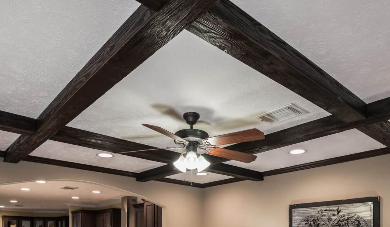 Ceiling with dark wooden beams forms a grid pattern. A ceiling fan with wooden blades is centered, surrounded by recessed lights, creating a cozy atmosphere.