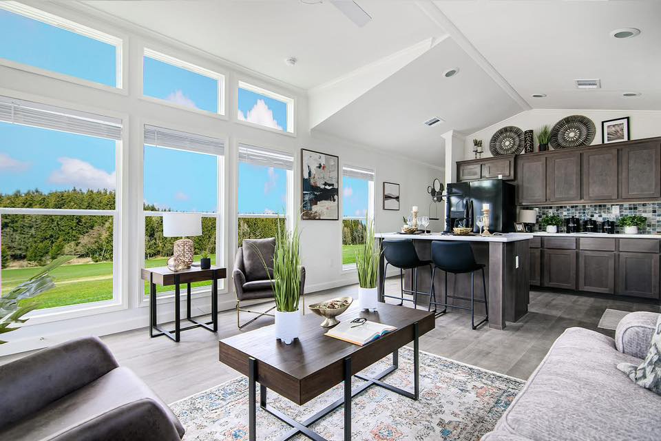 Bright, modern living room and kitchen with large windows showing a scenic outdoor view. Neutral tones, sleek furniture, and decor create a cozy feel.