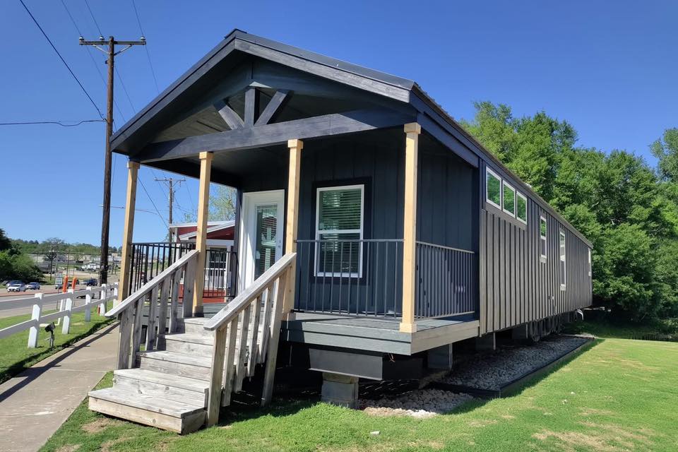 A modern tiny house with dark siding and light wooden accents sits elevated on a grassy lawn. Wooden steps lead to the front porch, bordered by railings. The scene is tranquil under a clear blue sky, suggesting a peaceful and inviting atmosphere.
