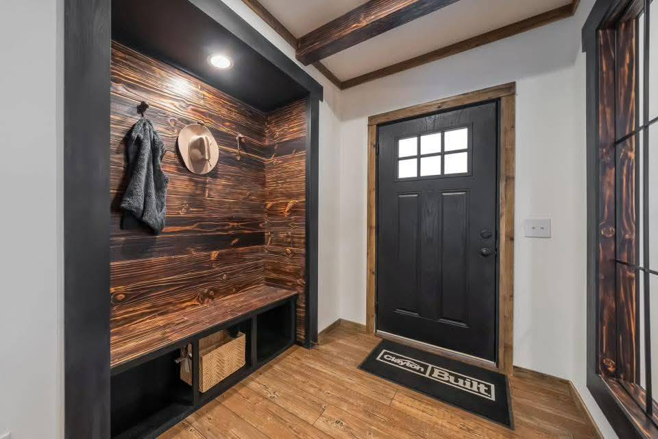 Rustic entryway with a black door, wood accents, and ceiling beams. A bench with storage below holds a basket. A coat and hat hang on hooks. Cozy and inviting.