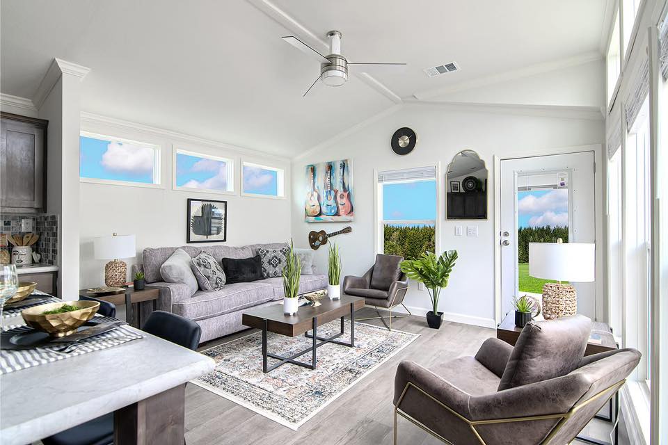 Bright living room with gray sofa, armchairs, and a coffee table on a patterned rug. Walls feature guitar art and windows offer a lush outdoor view.