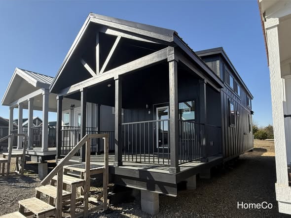 A modern tiny house with a dark wood exterior, covered porch, and metal railing. It sits elevated with front steps. Clear, sunny weather enhances its sleek look.