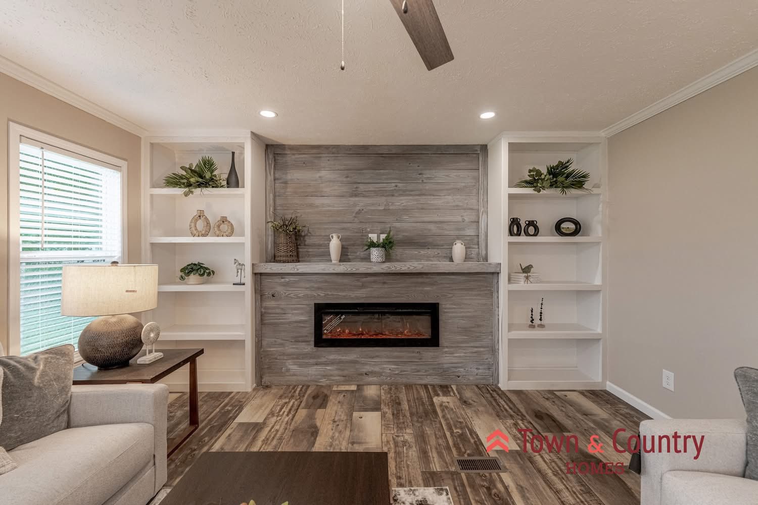 Cozy living room with a modern electric fireplace set in a wooden accent wall. Shelves hold plants and decor. Soft lighting and neutral tones create warmth.