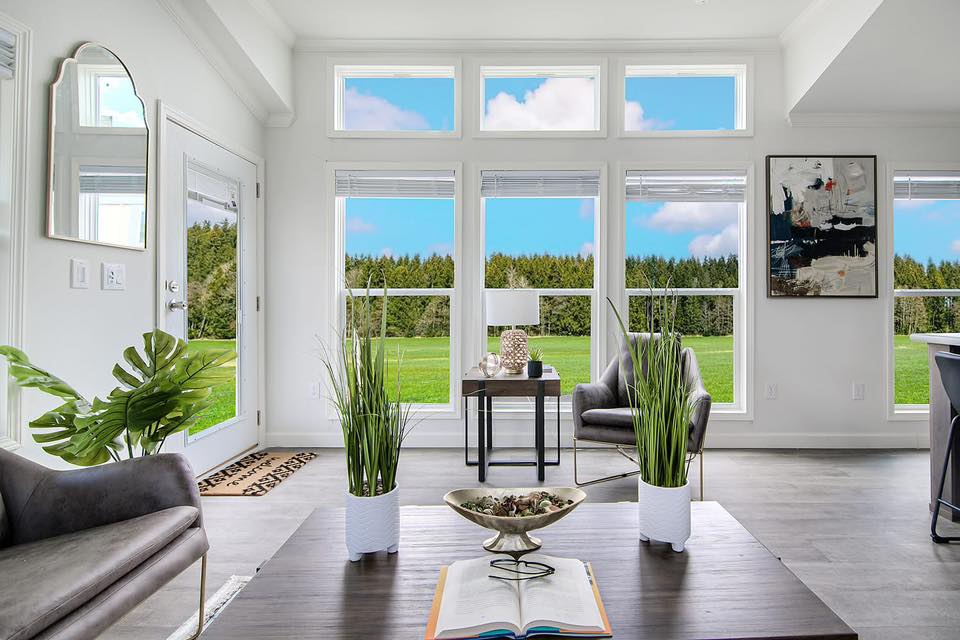 Bright living room with large windows showing a green field and blue sky. Modern decor includes plants, abstract art, and cozy seating. Inviting and tranquil.