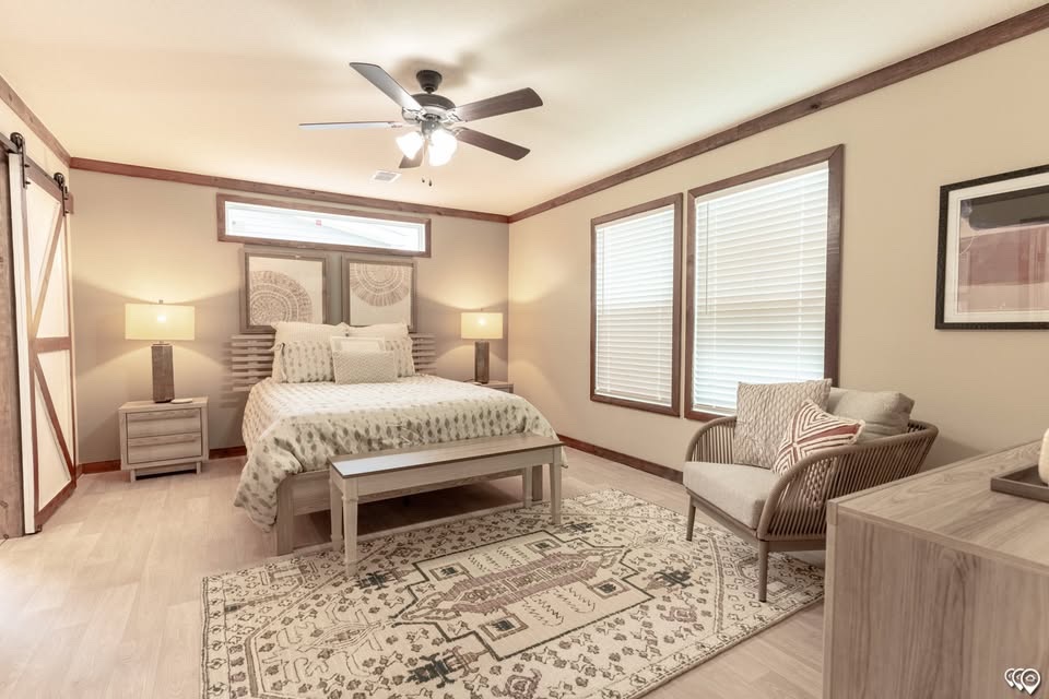 A cozy bedroom with a beige and wooden theme, featuring a double bed with patterned bedding, a rug, and a ceiling fan. Soft lighting and armchair create a warm atmosphere.