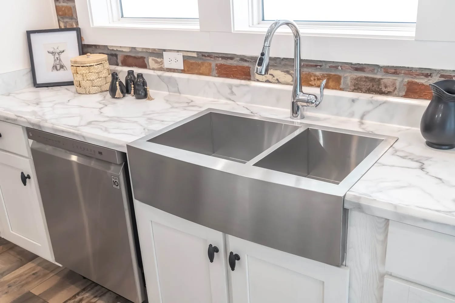 Modern kitchen scene with a stainless steel farmhouse sink, marble countertops, and white cabinetry. Decorative art and vases add a rustic touch.