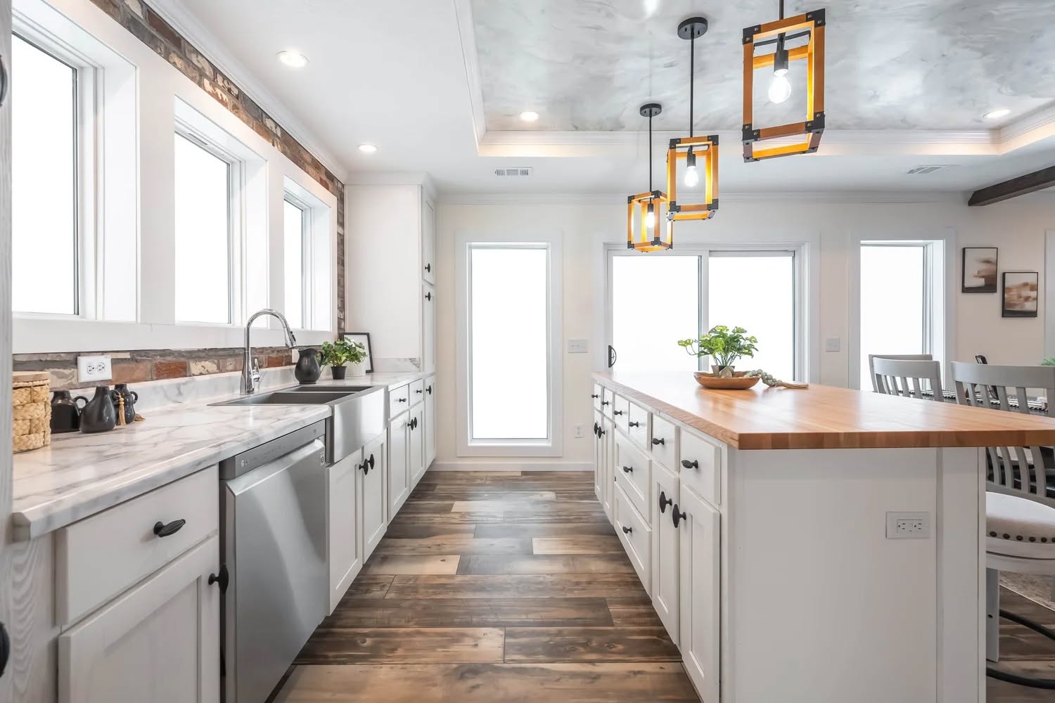 A bright, modern kitchen with white cabinets and wood countertops. Three pendant lights hang above a large island. Natural light streams through large windows.