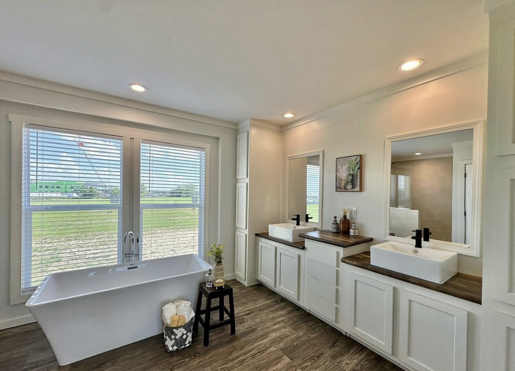 A modern bathroom with a freestanding tub and two square sinks on wooden vanities. Bright natural light enters through large windows, highlighting the elegant, serene design.