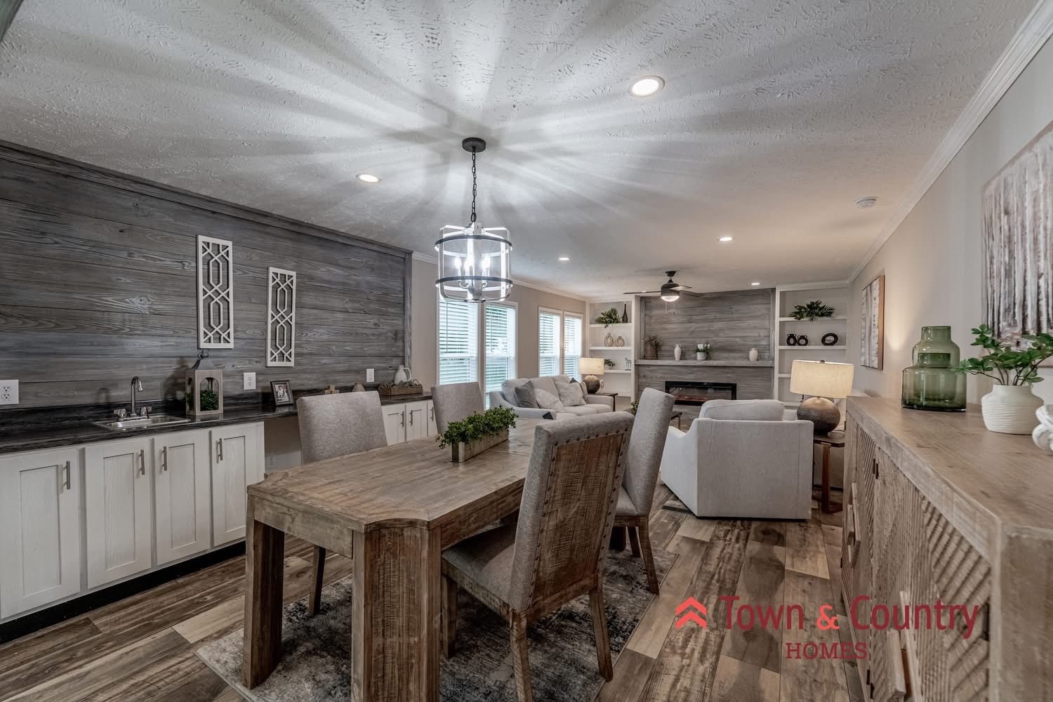Modern living and dining area with rustic charm. Wooden dining table, cushioned chairs, soft gray couches, and a cozy, decorative fireplace. Warm, inviting ambiance.