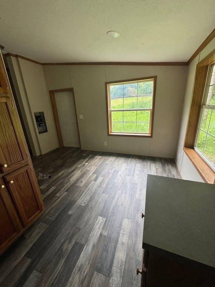 A small, empty room with wood laminate flooring, beige walls, and a large window. The window overlooks a grassy field, creating a serene atmosphere.