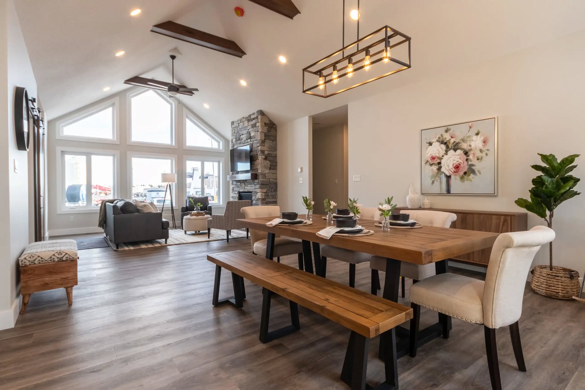 Spacious living-dining area with vaulted ceiling, large windows, and modern decor. Wooden dining table, gray sofas, and stone fireplace create a cozy ambiance.