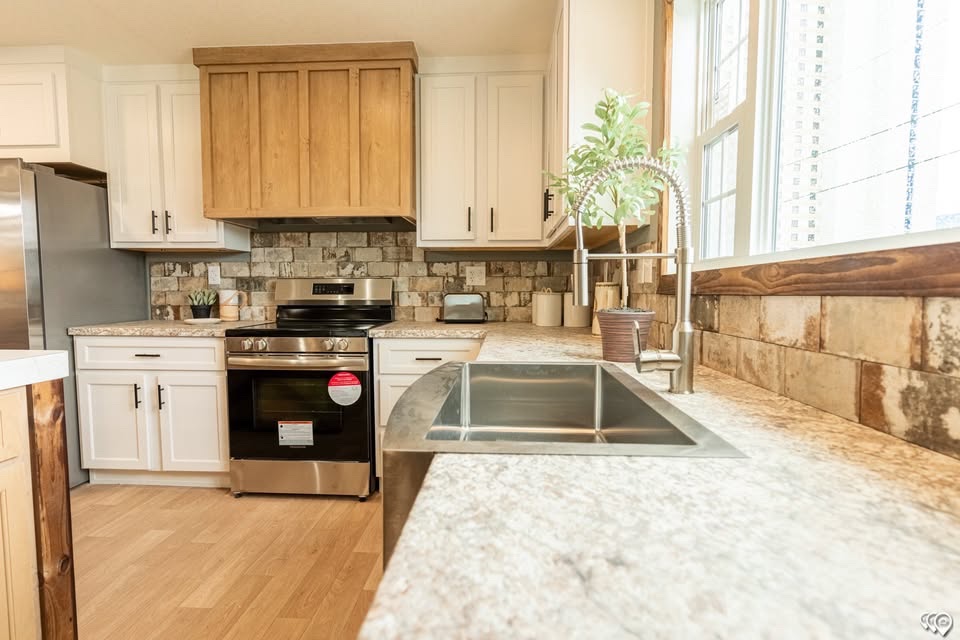 Modern kitchen with granite countertops, white and wood cabinets, stainless steel appliances, and a double sink. Bright light from large windows.